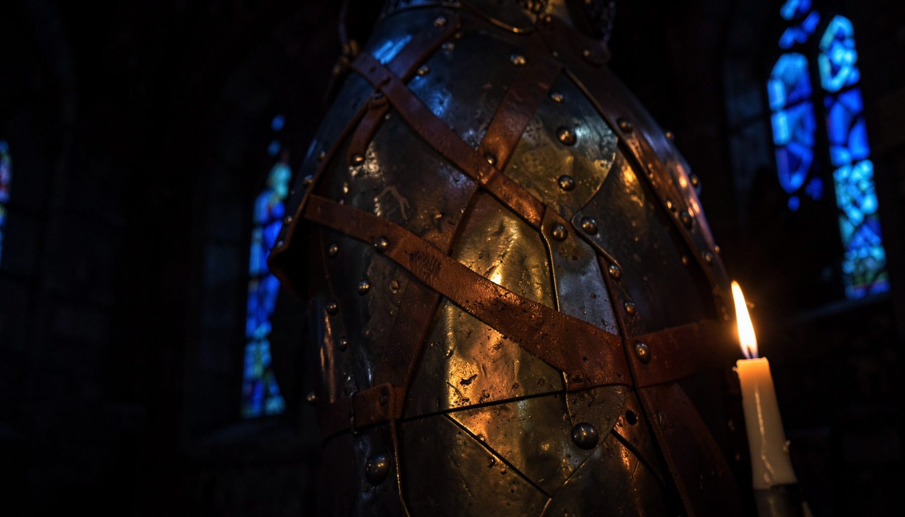 A moody close-up of riveted steel plate armour with leather straps, warm candlelight catching the metal's edge against a dark medieval interior.